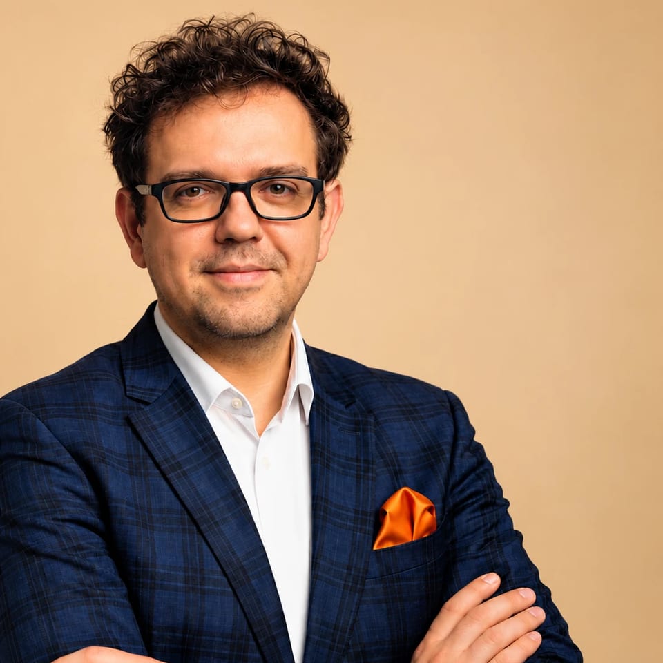 Paul Mardo w okularach i granatowej marynarce w kratę z pomarańczową poszetką, na beżowym tle